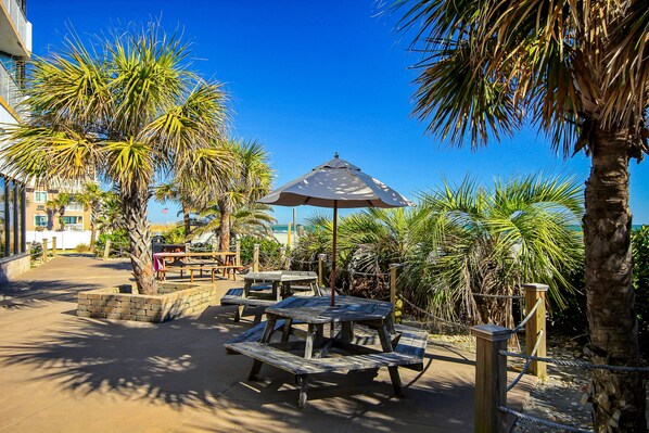 A patio area with picnic tables and an umbrella, surrounded by palm trees and a building in the background.