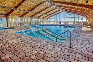 An indoor swimming pool with a brick floor and wooden ceiling, surrounded by tables and chairs.