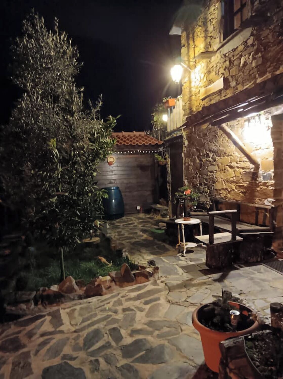 A stone patio with outdoor seating, a large tree, and a building with stone walls and a tiled roof.