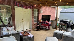 Une terrasse couverte, aménagée avec des meubles en osier, un barbecue et une télévision posée sur une table.