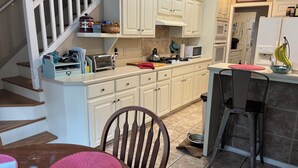 Une cuisine avec des armoires blanches, un four à micro-ondes, un grille-pain et un évier. On y trouve une table à manger en bois, avec des chaises et un set de table rouge. Un escalier est visible sur la gauche.