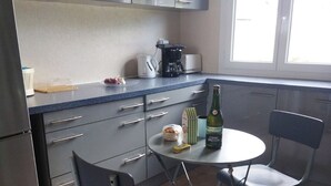 Eine kleine Küche mit grauen Schränken, einer Kaffeemaschine, einer Flasche Cidre Breton und zwei Stühlen um einen kleinen Tisch.