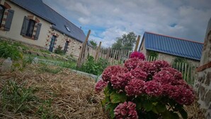 A rustic stone building with a sloped roof, surrounded by a wooden fence and a garden with hydrangea flowers. - Chambre de Charme, Gîte de L'archerie (Saint-Lambert-la-Potherie)