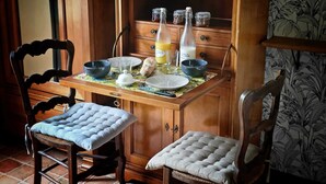 A wooden dining table with two chairs, a floral tablecloth, and a cabinet with glass jars and bottles. - Chambre de Charme, Gîte de L'archerie (Saint-Lambert-la-Potherie)
