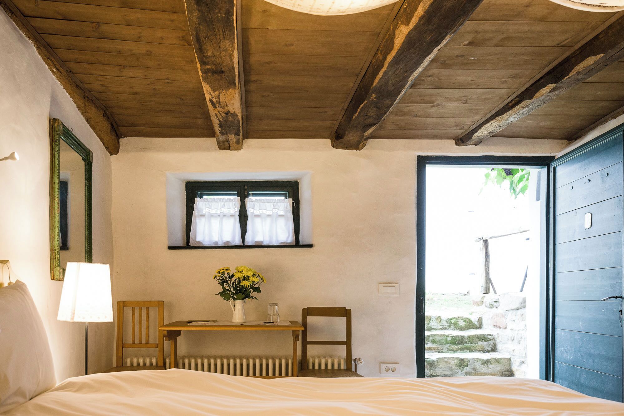 A bedroom with a wooden ceiling, a small window with white curtains, a bedside table with a vase of flowers, a chair, and a bed.