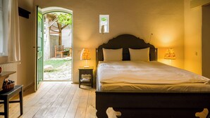 A bedroom with a wooden bed, two bedside lamps, a nightstand, and a view of a garden through an open door. - Viscri 125 (Bunesti)