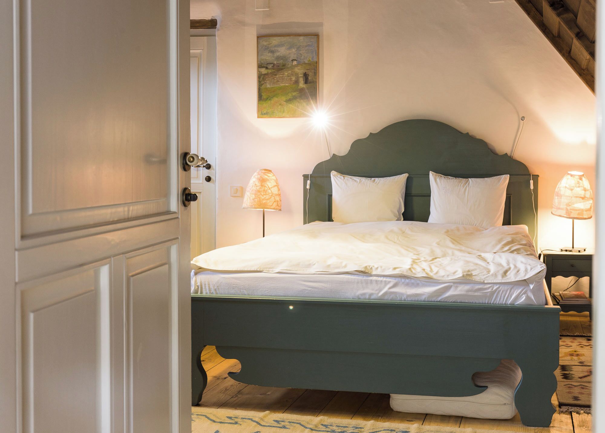 Ein Schlafzimmer mit einem Bett, zwei Nachttischlampen, einem Bild an der Wand und einer Tür.