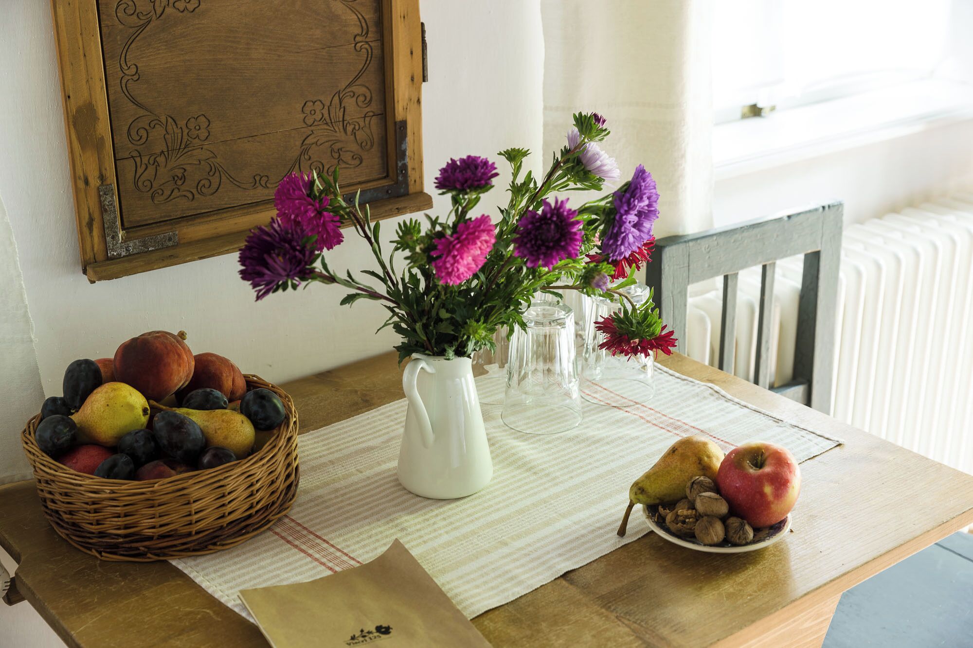 A table with a wicker basket of fruits, a vase with flowers, and a plate with nuts and a pear.