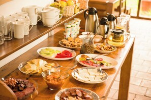 Un bufé de desayuno con diversos alimentos, como frutas, quesos y embutidos, además de una estación de café con varias cafeteras.