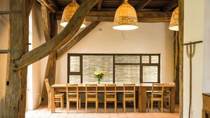 A rustic dining area with a wooden table and chairs, a vase of flowers, and a large window with blinds. - Viscri 125 (Bunesti)