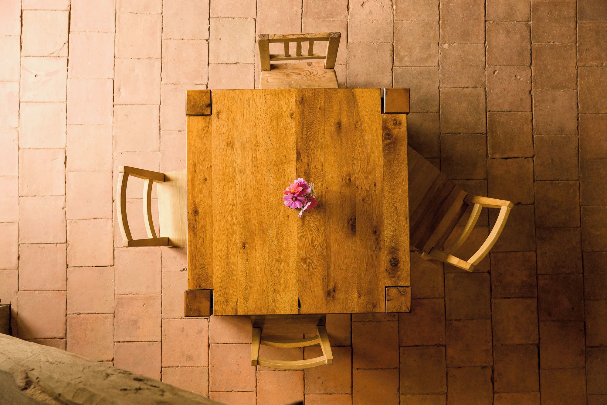 Una mesa cuadrada de madera con un centro de flores, rodeada por cuatro sillas de madera, frente a una pared de ladrillo.