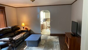 A living room with a leather sofa, a blue ottoman, a television on a wooden stand, and a lamp on a side table. - Quaint, private setting, 3-4 miles from SOMC & KDMC hosp. (Portsmouth)