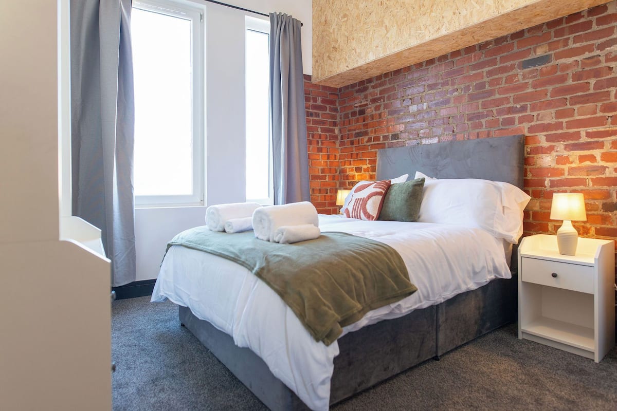 A hotel room with a brick wall, a large bed with white bedding, a nightstand with a lamp, and a window with curtains.