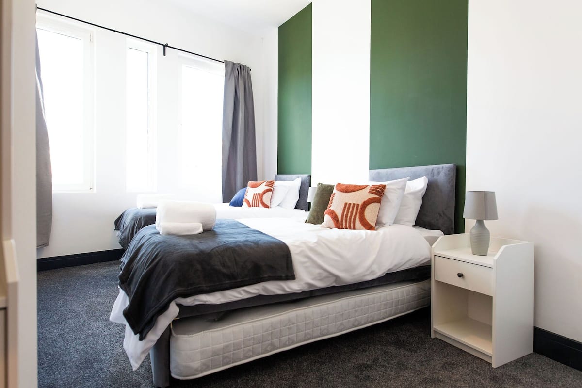 A neatly made bed with a white and dark gray duvet, a bedside table with a lamp, and a view of a window with curtains.