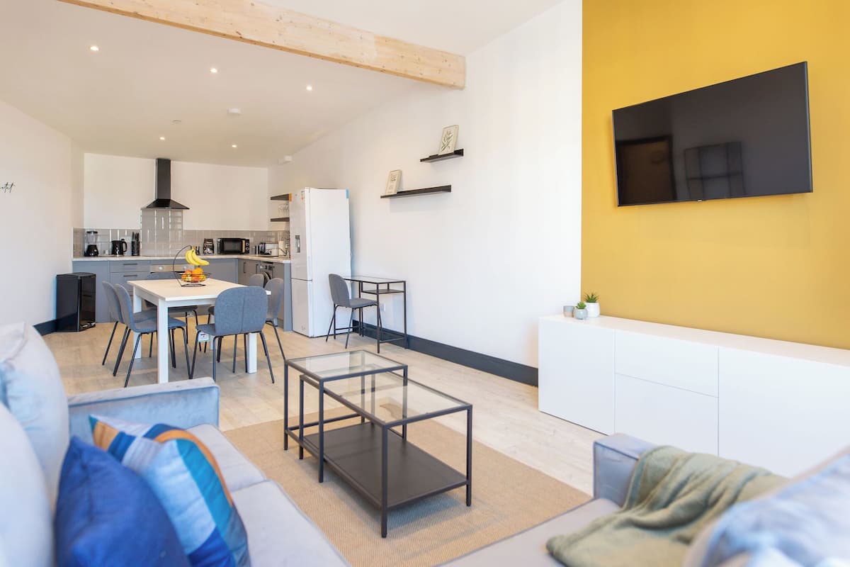 A modern kitchen with a dining area, a living room with a sofa and a coffee table, and a TV mounted on a yellow wall.