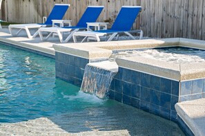 Une piscine agrémentée d’une cascade, des chaises longues bleues et une clôture en bois en arrière-plan.