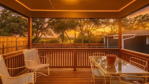 A covered outdoor patio with wooden flooring, wicker chairs, a glass table, and a view of trees and a sunset. - Wilbarger Villa-Sunsets Overlooking SWU & Downtown (Georgetown)