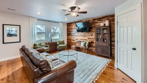 A living room with a leather sofa, a coffee table, and a TV mounted on a wooden wall. There is a ceiling fan, a bookshelf, and a door visible. - Wilbarger Villa-Sunsets Overlooking SWU & Downtown (Georgetown)