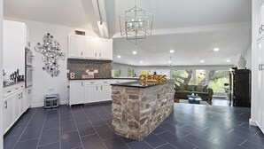 A modern kitchen with white cabinets, a stone island, and a dining area with a green sofa. - Your OASIS Nestled In Wooded River Views, POOL (Manchaca)