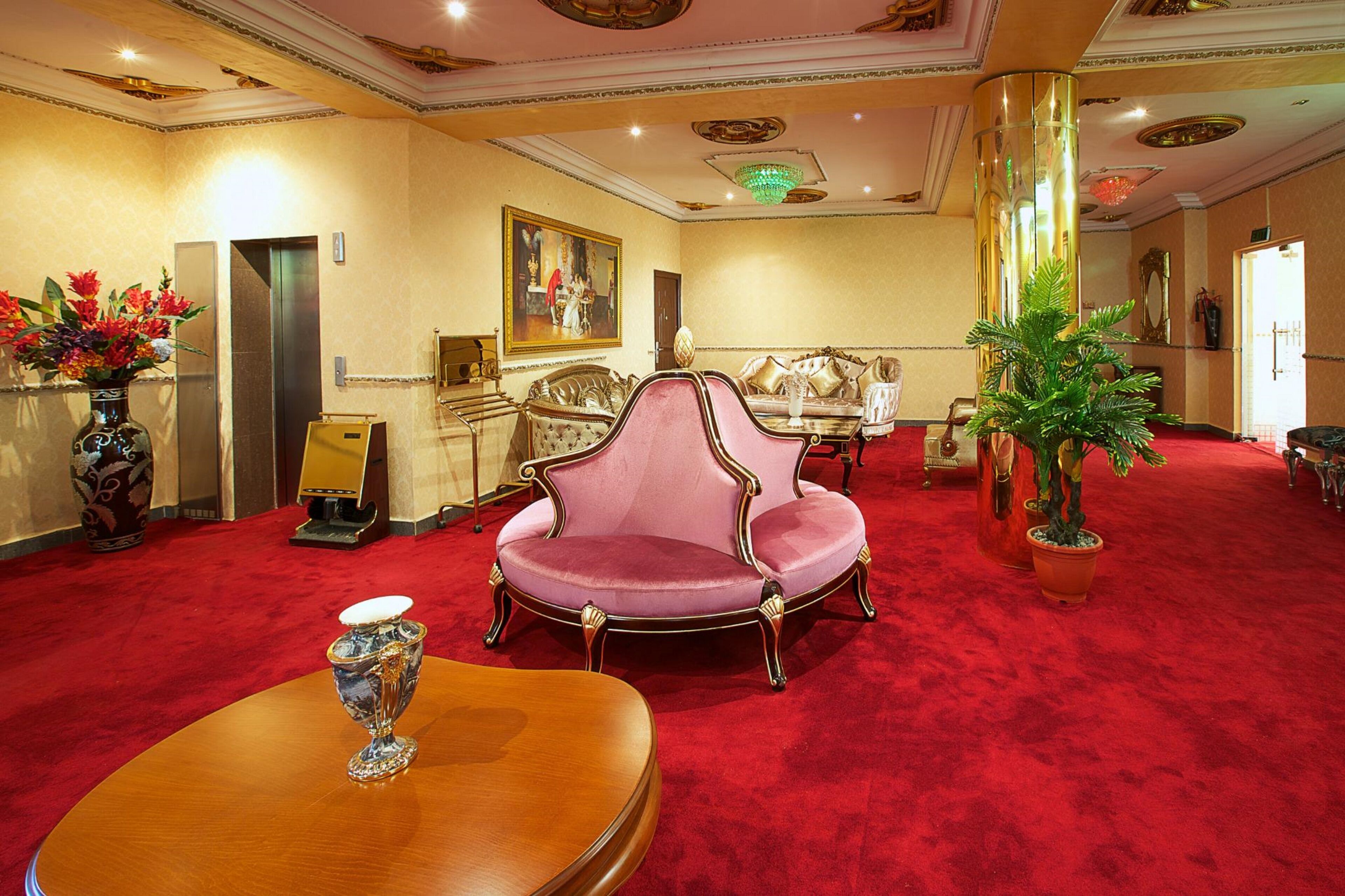 A lobby with a red carpet, a pink armchair, a wooden table with a vase, and a potted plant.