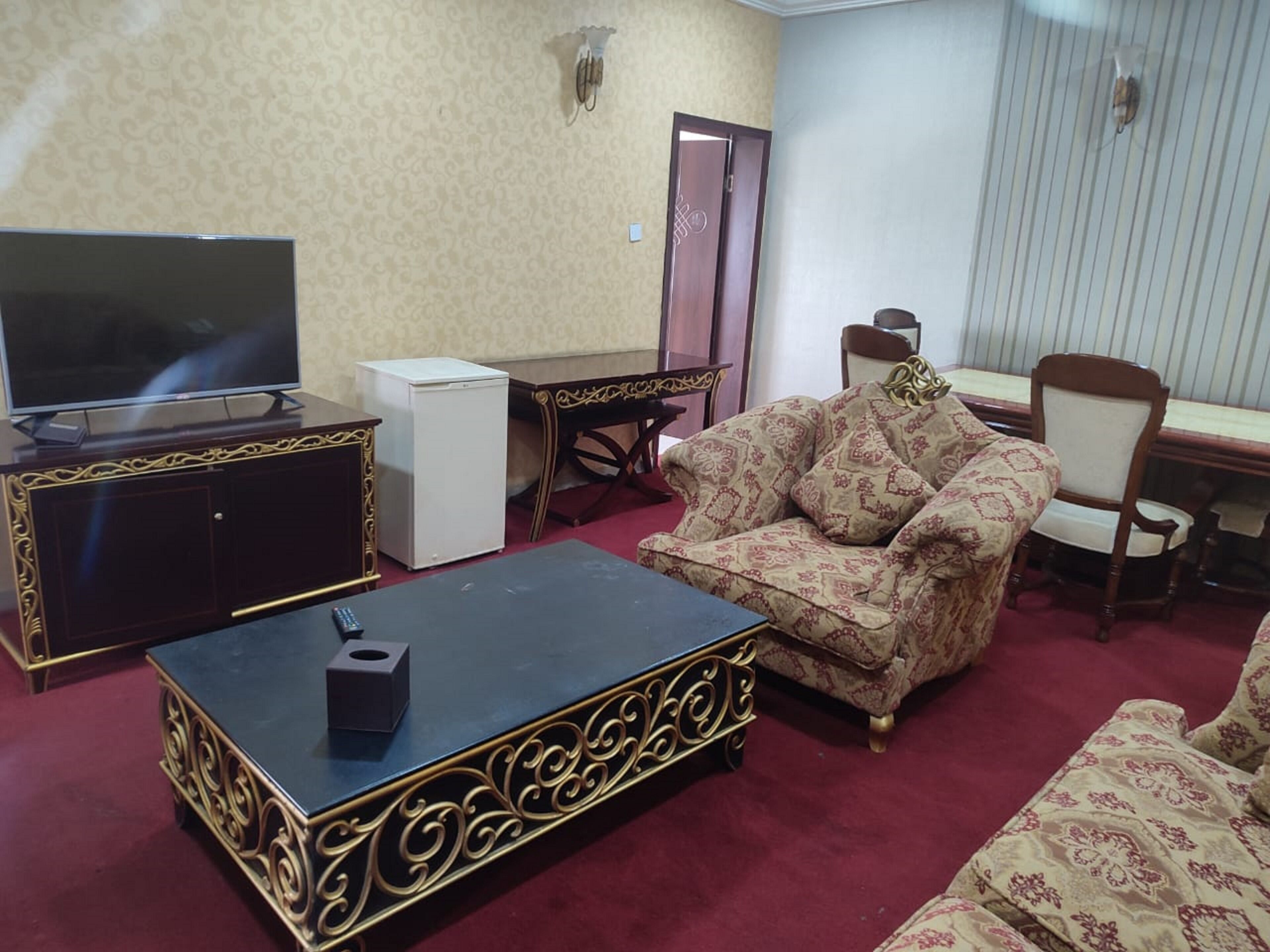 A living room with a TV, a small fridge, a patterned sofa, a coffee table, and a dining area with a chair and table.