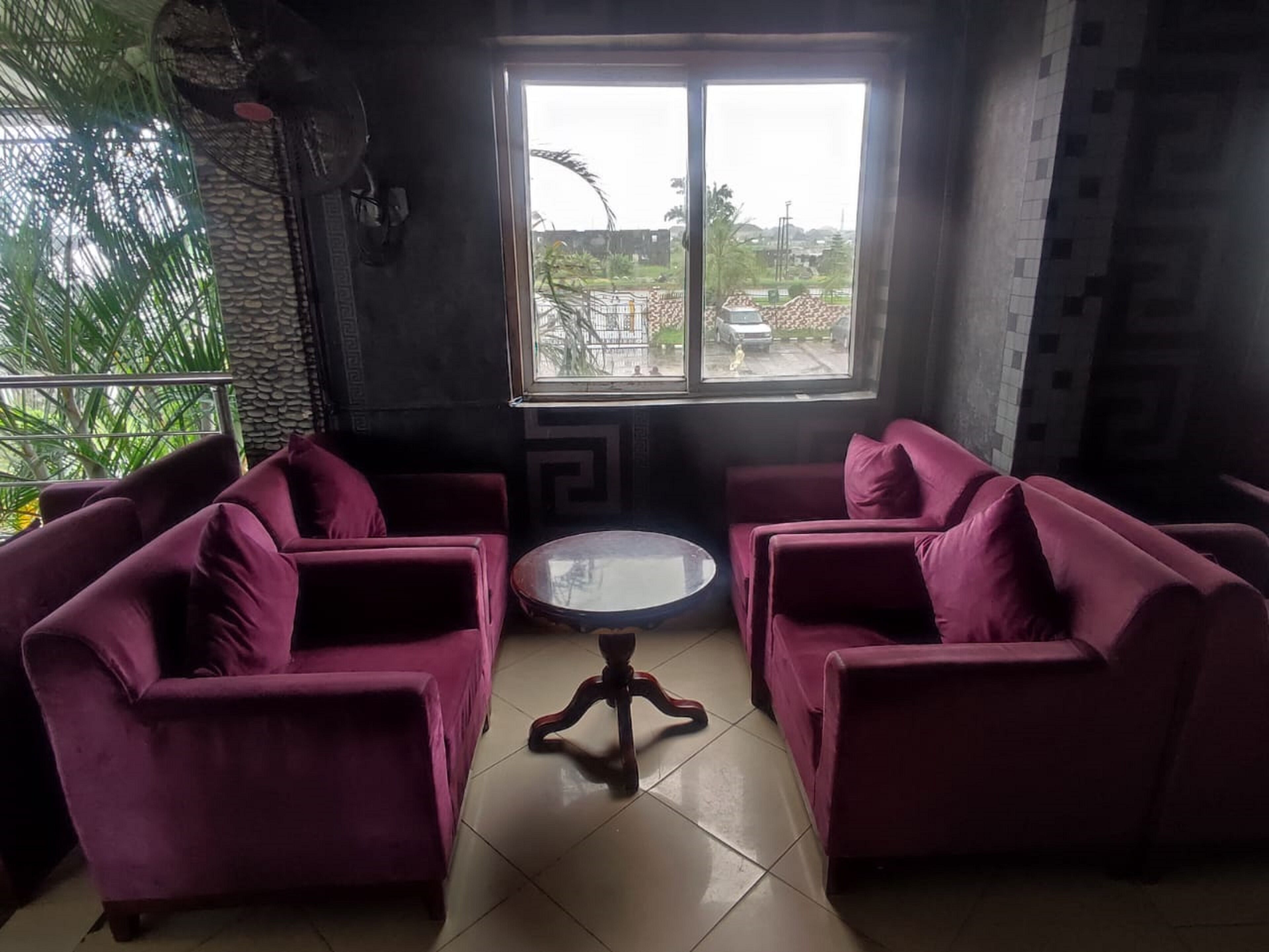 Une pièce avec deux canapés violets et une petite table ronde, une grande fenêtre donnant sur un paysage verdoyant et des bâtiments, et un mur décoratif orné d’un motif géométrique.