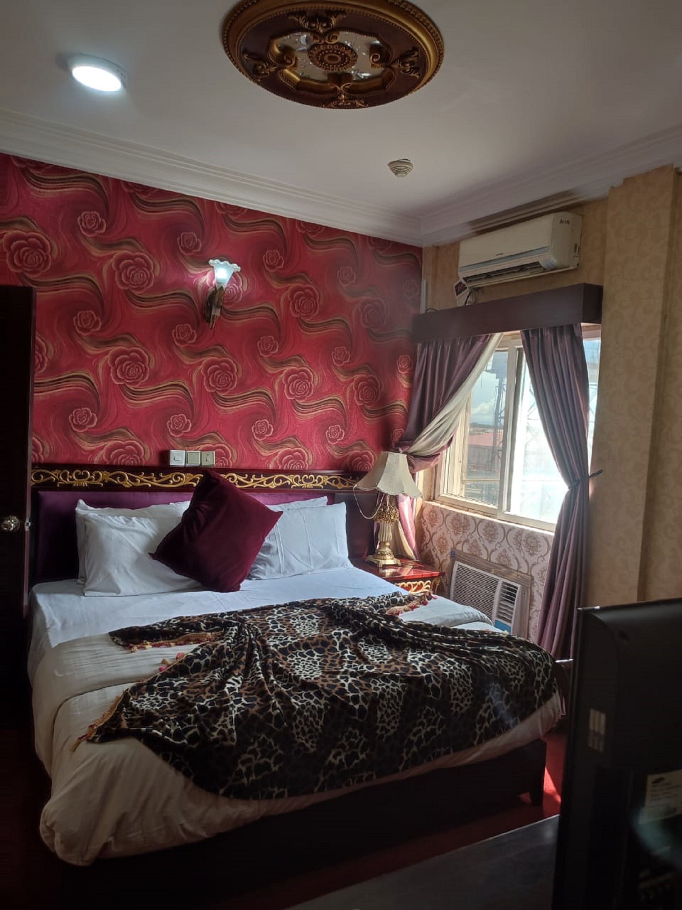 A hotel room with a large bed, red patterned wallpaper, a ceiling light, and an air conditioning unit.