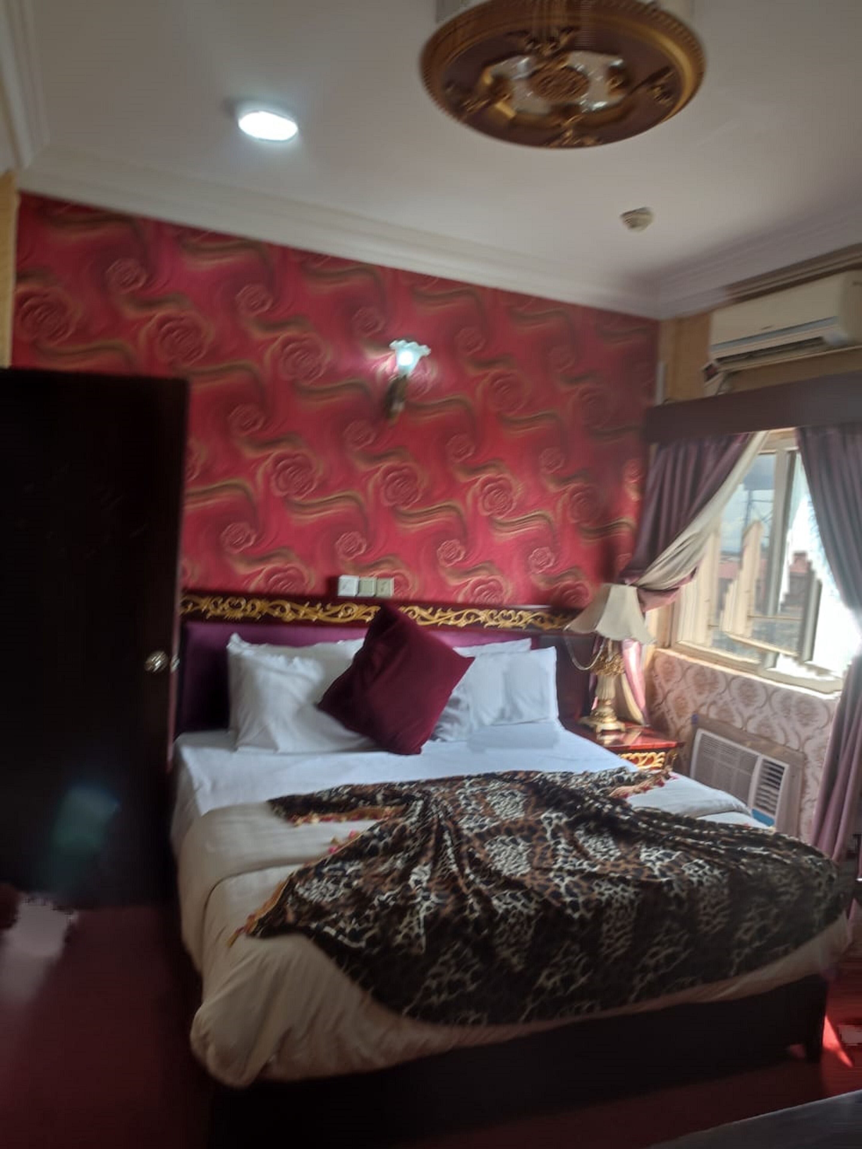 Una habitación de hotel con una cama grande, papel tapiz rojo con estampado y una ventana con cortinas.