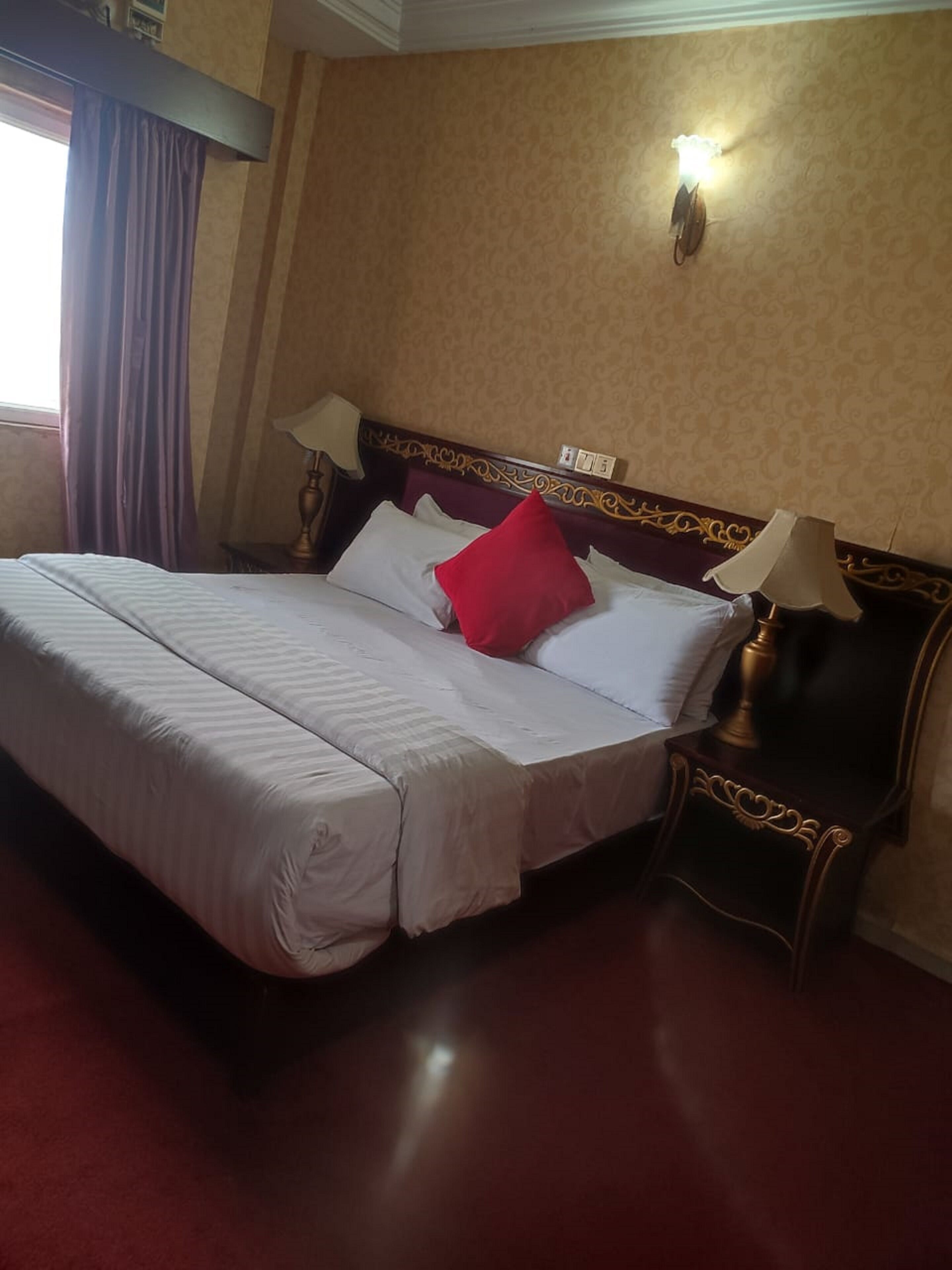 A neatly made bed with white linens and a red pillow, a wooden headboard with intricate carvings, a nightstand with a lamp, and a wall-mounted light fixture.