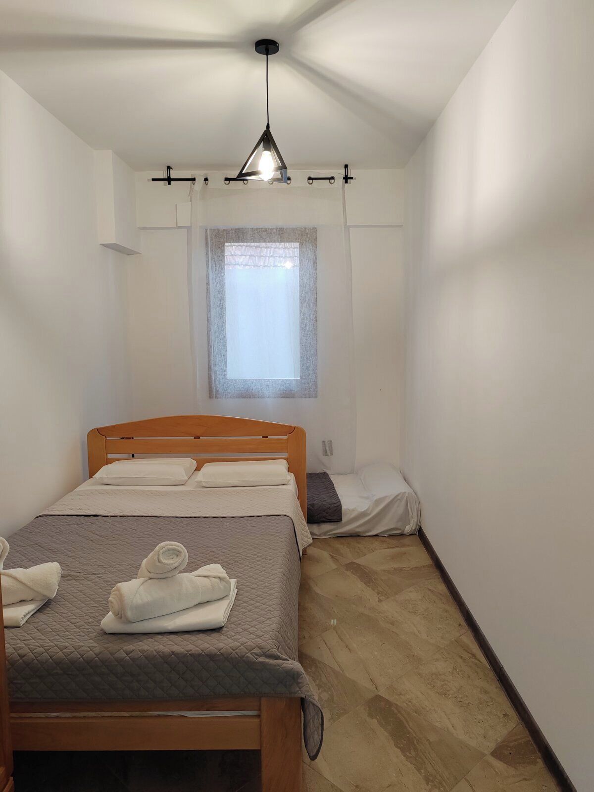 Una habitación pequeña y limpia, con una cama individual, una ventana con cortina translúcida y una lámpara colgante.