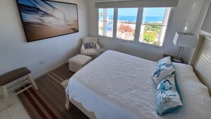 Un dormitorio con una cama grande, una silla, una mesita, un cuadro y una ventana con vistas.