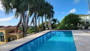 Una piscina con sillones de descanso, rodeada de palmeras y un edificio con techo de tejas.