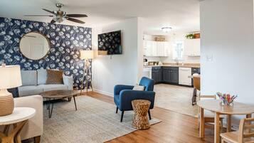 A modern living room with a sofa, coffee table, and a blue armchair. There is a kitchen in the background with white cabinets and a dining area with a table and chairs.