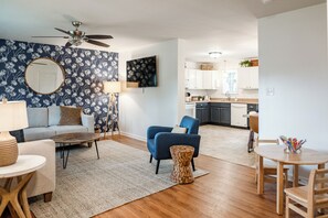 Ein modernes Wohnzimmer mit einer Couch, einem Couchtisch und einem blauen Sessel. Im Hintergrund befindet sich eine Küche mit weißen Schränken und ein Essbereich mit Tisch und Stühlen.