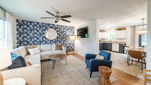 A modern living room with a sofa, coffee table, and blue armchair. There is a kitchen area with a dining table and chairs.