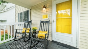 A porch with two rocking chairs, a small table with a potted plant, and a yellow door.
