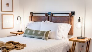 A neatly made bed with white linens, a green patterned pillow, and a brown throw blanket. There are two bedside tables with lamps, and a power outlet on the wall.