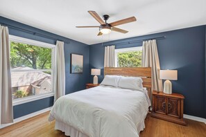 Un dormitorio con una cama, dos mesitas de noche con lámparas, un ventilador de techo y una ventana con cortinas.