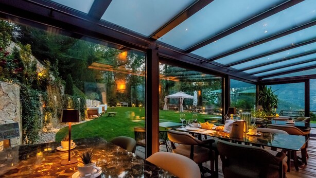A modern outdoor dining area with a glass ceiling, a stone wall with greenery, and a view of a well-maintained lawn and garden. - Chalet Bragard - Boutique Chalet & Spa (Limone Piemonte)