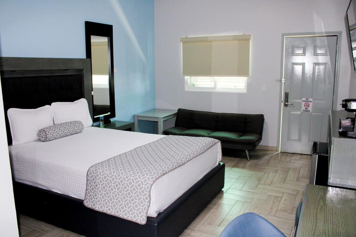 Une chambre d’hôtel comprenant un lit, un bureau, une chaise, un canapé, un miroir, une fenêtre avec des stores et une porte sur laquelle est apposé un panneau « interdiction de fumer ».