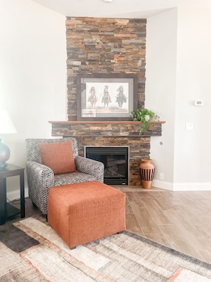A living room with a stone fireplace, a framed picture above it, a patterned armchair, an ottoman, and a rug.