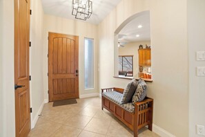A hallway with a wooden door, a bench with patterned cushions, a mirror, and a window with blinds.