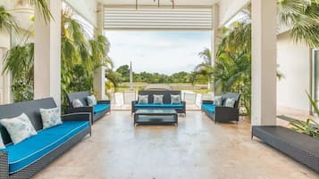 A covered outdoor seating area with sofas and a coffee table, surrounded by palm trees and a view of the landscape.