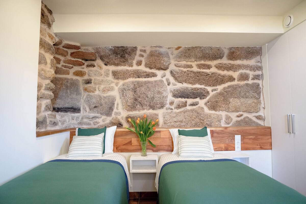 A room with two beds, a stone wall, a wooden headboard, and a vase with flowers.