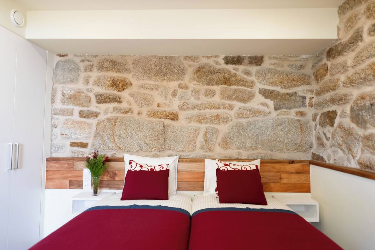 A bedroom with a stone wall, wooden headboard, two beds with red bedding, a nightstand with a vase of flowers, and a white wardrobe.