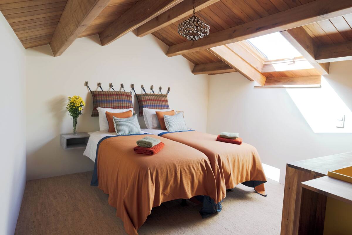 A bedroom with two beds, wooden beams, a skylight, and a hanging light fixture.