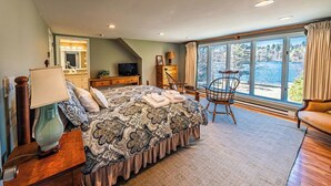 A bedroom with a large bed, a TV, a dresser, and a chair by a window overlooking a body of water. - Lake Cottage | 5 Bed, 5 Bath (Lake Placid)