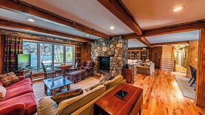 A cozy living room with a stone fireplace, wooden beams, and a large window overlooking a scenic view. - Lake Cottage | 5 Bed, 5 Bath (Lake Placid)