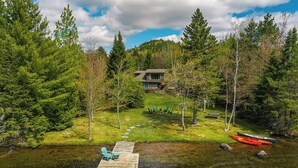 A lakeside cabin with a dock, surrounded by dense forest and a clear sky. - Lake Cottage | 5 Bed, 5 Bath (Lake Placid)