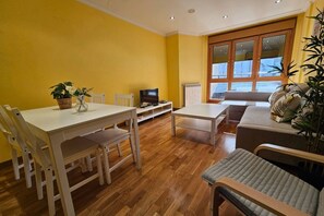 Een woonkamer met een eettafel, stoelen, een televisie, een bank en een raam met zonwering.